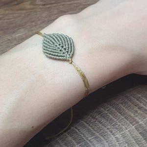 Könnte beinhalten: Ein hellgrünes Makramee-Armband mit einem blattartigen Design. Das Armband ist mit einer goldenen Schnur gebunden.