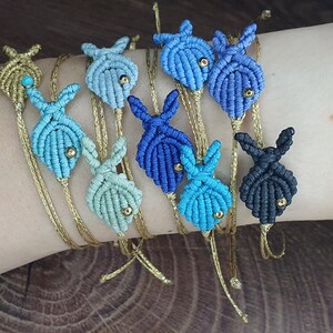 Peut inclure: Collection de bracelets en macramé avec des breloques en forme de poisson dans différentes nuances de bleu et d'or. Chaque breloque a une petite perle dorée. Les bracelets sont présentés sur un poignet.