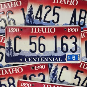 May include: Vintage Idaho license plates with a red and white gradient background. The plates feature black lettering and numbers, with a mountain and pine tree graphic. The text "IDAHO" and "CENTENNIAL" are visible.