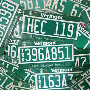 Peut inclure: Gros plan sur une pile de plaques d'immatriculation vertes du Vermont avec des lettres blanches. Les plaques comportent le nom de l'État, "Green Mountain State", et divers numéros de plaques d'immatriculation, notamment "HEC 119", "396A851" et "163A".