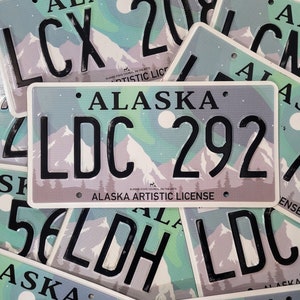 Könnte beinhalten: Nahaufnahme eines Stapels von künstlerischen Kennzeichen aus Alaska. Die Schilder sind weiß mit einer blauen und grünen Bergszene und dem Text "ALASKA ARTISTIC LICENSE". Die Kennzeichen-Nummer lautet "LDC 292".