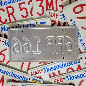 May include: A close-up of a silver license plate with the numbers "661996" embossed in black. The plate is surrounded by other license plates with various numbers and the words "Massachusetts" and "Spirit of America".