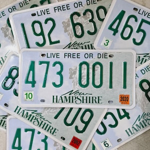 May include: A close-up of a stack of New Hampshire license plates with the state motto "Live Free or Die" printed on them. The plates are white with green numbers and text. The plates are from different years, including 2002 and 2020.