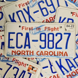 May include: A white North Carolina vehicle registration plate with blue embossed characters reading ECA-9827. The plate features the red text First in Flight, a blue graphic of an aeroplane, and a white registration sticker in the corner marked 04-22.