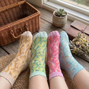 Op de afbeelding: Vier paar kleurrijke sokken met een semi-transparant ontwerp. De sokken zijn in tinten geel, groen, roze en blauw. De sokken liggen op een geweven mat, met een picknickmand en een kleine potplant op de achtergrond.