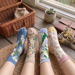 Op de afbeelding: Vier paar sokken met bloemmotieven. Eén paar is blauw met witte madeliefjes, een ander is beige met bruine bloemen, een derde is crème met paarse bloemen en de laatste is crème met roze bloemen. De sokken liggen op een geweven mat.