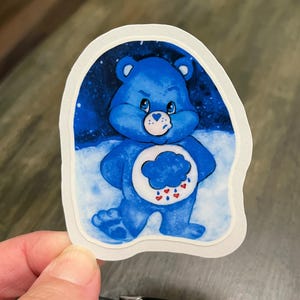 May include: A die-cut sticker featuring a blue bear character with a white belly patch displaying a blue cloud with red hearts and blue raindrops. The bear is set against a dark blue starry background and a white snowy foreground.