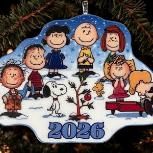 May include: A Peanuts-themed Christmas ornament featuring Charlie Brown, Snoopy, Woodstock, and friends gathered around a small tree in the snow. The characters are illustrated in a winter setting with the year 2026 printed in blue at the bottom.