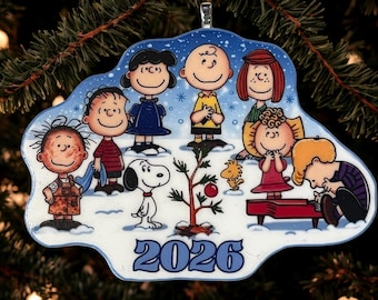 May include: A Peanuts-themed Christmas ornament featuring Charlie Brown, Snoopy, Woodstock, and friends gathered around a small tree in the snow. The characters are illustrated in a winter setting with the year 2026 printed in blue at the bottom.