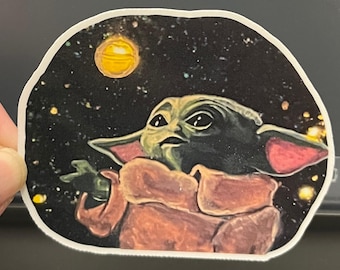 May include: A die-cut sticker featuring a green, large-eared creature with dark eyes wearing a tan robe. The character is set against a dark, starry space background with a bright yellow planet or moon visible in the upper left corner of the design.