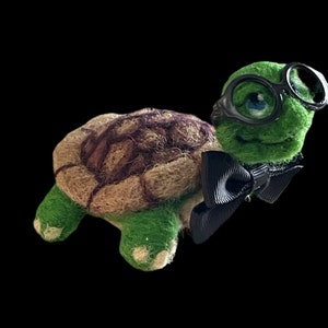 May include: A green and brown felt turtle wearing black-rimmed glasses and a black bow tie.