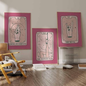 May include: Three rectangular wall art prints featuring tarot card designs in shades of pink and dark green. The left print shows a hand with fingers crossed labeled Wheel of Fortune, the center shows a leg hanging labeled The Hanged Man, and the right shows a hand gesture labeled The Devil.