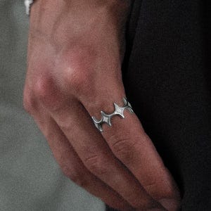 Könnte beinhalten: Ein silberner Ring mit sich wiederholendem Sternmuster wird an einer Hand getragen. Ein passendes silbernes Armband ist am Handgelenk zu sehen. Der Hintergrund ist ein neutrales Grau, mit einem schwarzen Kleidungsstück auf der rechten Seite.