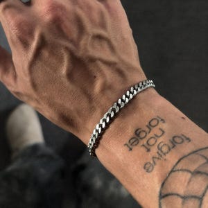 Könnte beinhalten: Ein silbernes Kettenarmband und ein Ring mit einem Panzerketten-Design. Der Ring befindet sich an einem Finger und das Armband an einem Handgelenk. Das Handgelenk hat ein Tattoo mit der Aufschrift "Forgive not forget".