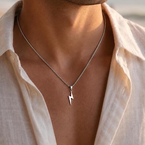 Könnte beinhalten: Silberfarbene Halskette mit einem Blitz-Anhänger. Die Halskette wird über einem hellen Hemd mit Knöpfen getragen. Die Kette hat ein strukturiertes Design. Der Anhänger ist schlicht und minimalistisch gestaltet.