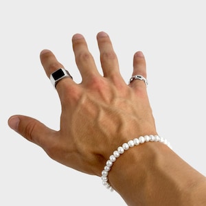Könnte beinhalten: Eine Hand mit einem Perlenarmband und zwei Silberringen. Ein Ring hat einen schwarzen quadratischen Stein, der andere ein Kettendesign. Das Armband besteht aus kleinen, runden, weißen Perlen.