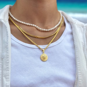 Könnte beinhalten: Ein mehrschichtiges Halsketten-Set mit einer Perlenkette, einer Goldkette und einem Goldanhänger mit detailliertem Design. Die Halsketten werden über einem weißen Rippen-Tanktop und einem weißen Hemd mit Knöpfen getragen.