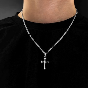 Könnte beinhalten: Eine silberne Kettenkette mit einem silbernen Kreuzanhänger. Das Kreuz hat ein schlichtes, klassisches Design.