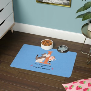 May include: Blue pet mat with a cartoon cat and a boy sharpening their claws on a scratching post. The mat has the text "Just sharpening my murder mittens!" printed on it.