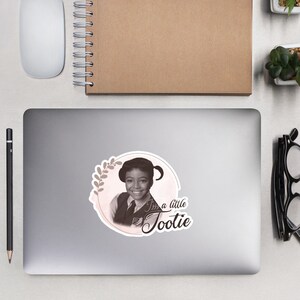 May include: A sticker featuring a black and white photo of a young girl with a floral wreath around her head. The text "I'm a little Tootie" is written in cursive below the photo.