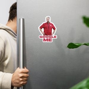 May include: A red magnet featuring a man in a red shirt with the text "GOOGLE ME" in white letters. The magnet is affixed to a silver refrigerator door.