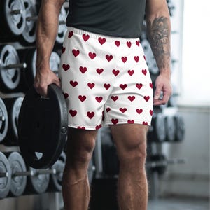 May include: Athletic shorts featuring a repeating pattern of small, solid dark red hearts on a white background. The shorts have an elastic waistband and a relaxed fit, designed for movement and exercise, showcasing a simple and clean aesthetic.