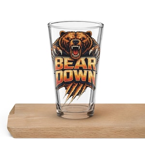 Könnte beinhalten: Ein klares Bierglas mit einem braunen Bärengrafik und dem Schriftzug "BEAR DOWN" in Orange und Gelb. Der Bär hat einen geöffneten Mund und Krallen. Das Glas steht auf einer Holzoberfläche.