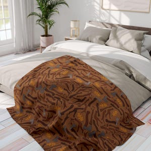 May include: A brown throw blanket with a repeating pattern of axes and leaves. The axes are various shades of brown and gray, and the leaves are yellow. The blanket is draped over a bed with neutral-colored bedding.