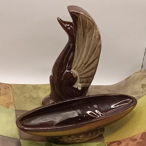 May include: A brown ceramic bird figurine with outstretched wings, perched above a matching oval dish. The bird has a detailed wing design and a glossy finish. The dish is supported by a decorative base.