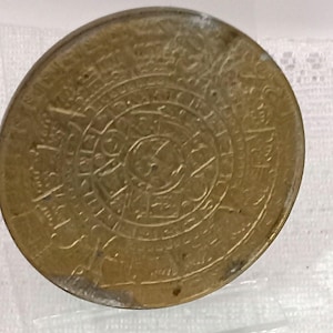 May include: A circular brass-colored metal medallion featuring intricate embossed concentric patterns and symbols. The surface displays a central circular motif surrounded by multiple rings of detailed engravings and geometric markings.