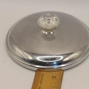 May include: A shiny, stainless steel lid with a clear, round knob. The lid is circular and has a slightly domed shape. A wooden ruler is placed next to the lid for scale.