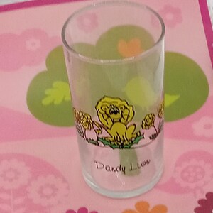 May include: A clear glass tumbler featuring a colorful graphic of a yellow lion surrounded by flowers. The text Dandy Lion is printed in black script below the illustration. The glass has a cylindrical shape with a smooth, transparent surface.