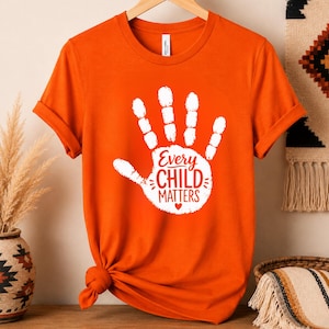 May include: A bright orange short-sleeved t-shirt featuring a large white handprint graphic on the center chest. Inside the handprint, the text reads Every CHILD MATTERS with a small heart symbol below the words, printed in a bold orange font.