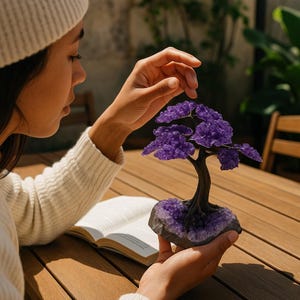 May include: A decorative amethyst tree sculpture with a dark brown trunk and vibrant purple leaves. The tree sits on a base of raw amethyst crystals. The sculpture is held in a person's hands, with a book open on a wooden table in the background.