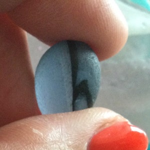Puede incluir: Una piedra lisa de forma ovalada con una franja gris oscura que recorre el centro. La piedra es de color azul claro.