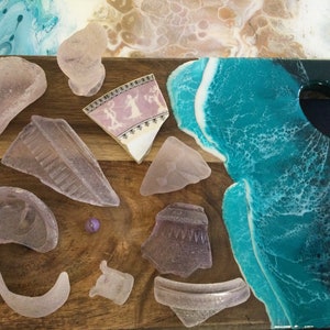 Könnte beinhalten: Ein Holztablett mit einem blauen und weißen Ozeanwellen-Design aus Harz. Das Tablett ist mit verschiedenen Stücken violettem Strandglas und einer kleinen violetten Perle verziert.