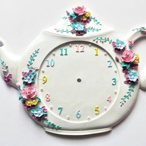 Könnte beinhalten: Weiße Teekannen-Wanduhr mit bunten floralen Akzenten. Das Zifferblatt hat Zahlen in verschiedenen Farben und ist mit rosa, blauen und gelben Blumen verziert. Die Uhr hat einen Griff und einen Ausguss.