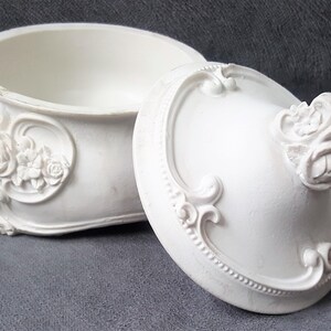 May include: A white, oval-shaped trinket box with an open lid. The box and lid are adorned with floral and scrollwork details. The lid features a prominent floral handle. The box is made of a matte material.