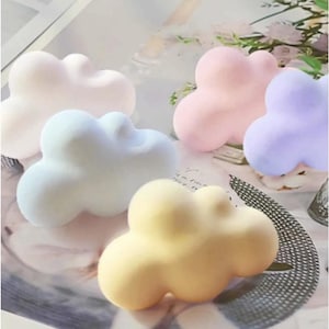 Puede incluir: Cinco objetos con forma de nube en colores pastel: blanco, azul claro, rosa, morado y amarillo. Los objetos son lisos y redondeados, con un aspecto suave y esponjoso. Las nubes están dispuestas sobre una superficie de color claro.