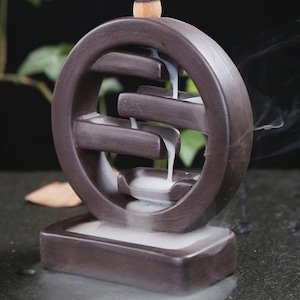 Könnte beinhalten: Ein brauner Keramik-Räuchergefäß mit kreisförmigem Design und kaskadierenden Ebenen. Weißer Rauch fließt die Ebenen hinunter und erzeugt einen Wasserfalleffekt. Der Brenner steht auf einem rechteckigen Sockel, mit einem angezündeten Räucherkegel oben.