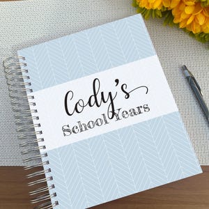 May include: A spiral-bound notebook with a light blue and white herringbone pattern cover. A central white horizontal band features the black text "Cody's School Years" in a combination of script and serif fonts. A black pen rests nearby.