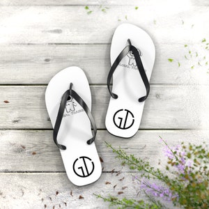 May include: White flip-flops with black straps, featuring a black and white logo of a cartoon astronaut and the letters "GD". The flip-flops are on a light gray wooden surface, with some greenery in the background.