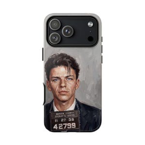 May include: A smartphone case featuring a painted portrait of a young man with dark hair and blue eyes, wearing a dark suit. The subject holds a Bergen County Sheriff's Office mugshot sign with the text 11 27 38 and 42799 printed in white.