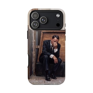 May include: A smartphone case featuring a printed image of a person in a dark suit and tie sitting on a dark trunk. The person has one hand covering their face and is positioned in a room with wooden panels. The case has a black camera cutout.