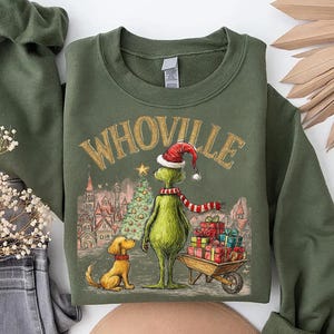 May include: Olive green sweatshirt featuring a graphic of a green character in a Santa hat and scarf, standing next to a dog and a cart of presents. The word "WHOVILLE" is printed above the character in gold lettering. A Christmas tree and buildings are in the background.