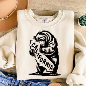 May include: A cream-colored sweatshirt with a black graphic of a bear hugging a California outline. The text "CALIFORNIA" is written inside the outline.