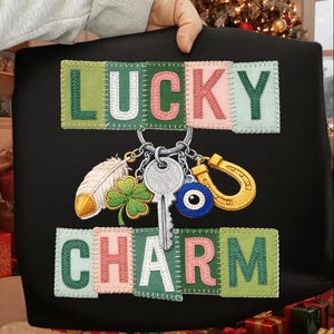 Könnte beinhalten: Schwarze Tragetasche mit den Worten "LUCKY CHARM" in gestickten Buchstaben. Die Tasche hat einen Schlüsselanhänger mit einem Feder-, Kleeblatt-, Schlüssel-, bösen Blick- und Hufeisen-Anhänger.