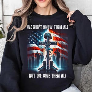 Op de afbeelding: Zwarte sweatshirt met een grafisch ontwerp met een kruis, geweer, laarzen en helm voor een Amerikaanse vlag. De tekst luidt: "WE DON'T KNOW THEM ALL BUT WE OWE THEM ALL".
