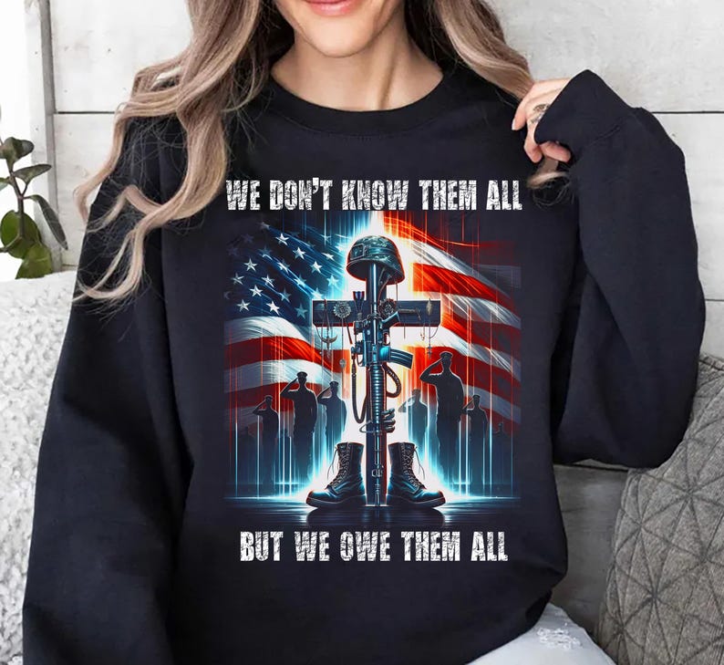 Op de afbeelding: Zwarte sweatshirt met een grafisch ontwerp met een kruis, geweer, laarzen en helm voor een Amerikaanse vlag. De tekst luidt: "WE DON'T KNOW THEM ALL BUT WE OWE THEM ALL".
