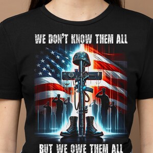 Op de afbeelding: Zwart T-shirt met een afbeelding van een kruis, geweer, helm en laarzen tegen een Amerikaanse vlag. De tekst luidt: "WE DON'T KNOW THEM ALL BUT WE OWE THEM ALL".